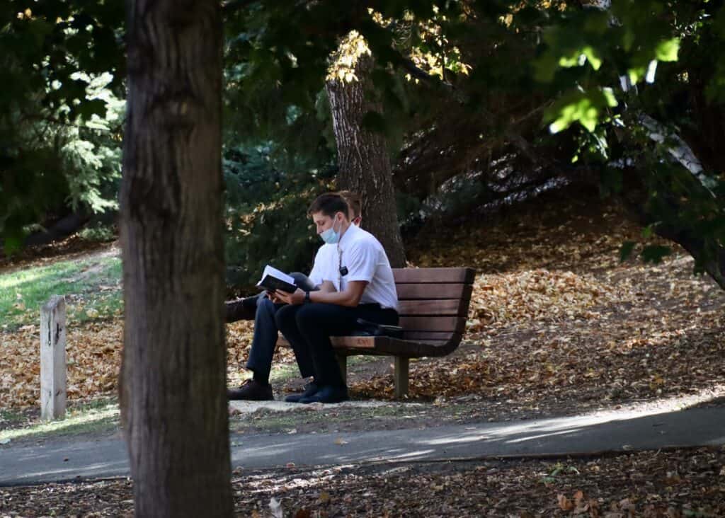 Beliefs - two missionaries on a park bench wearing masks and reading Scripture