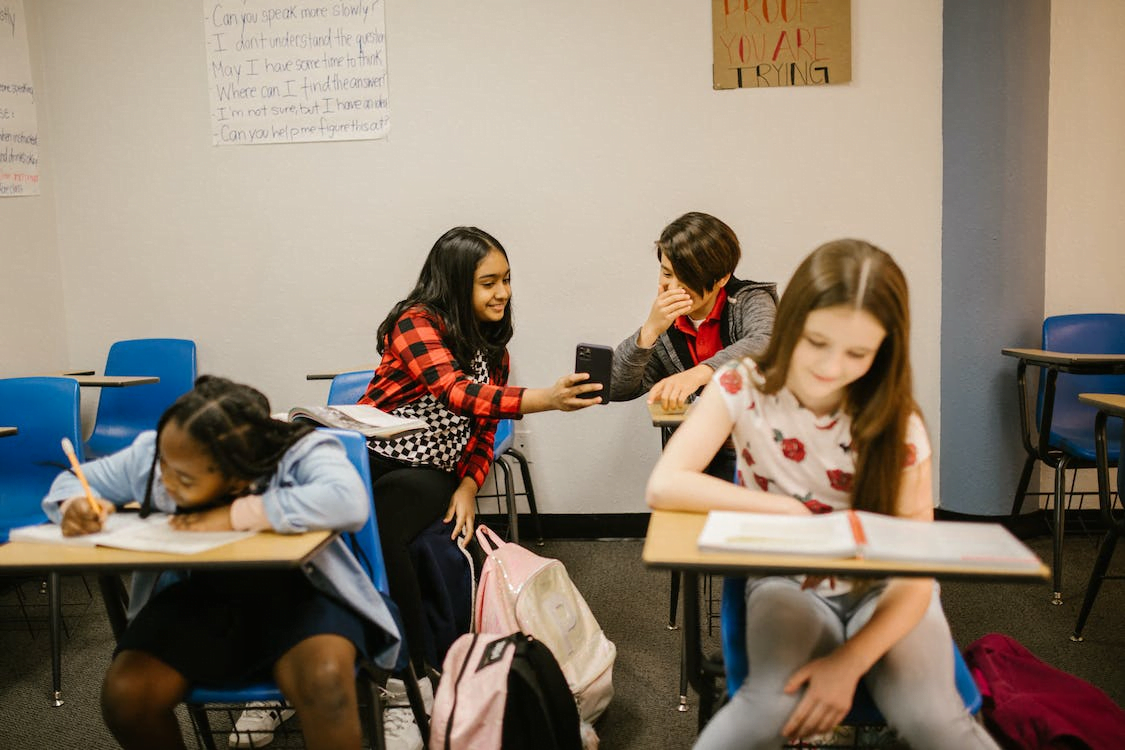 a group of children sitting at desks in a classroom, two children using a phone at the back