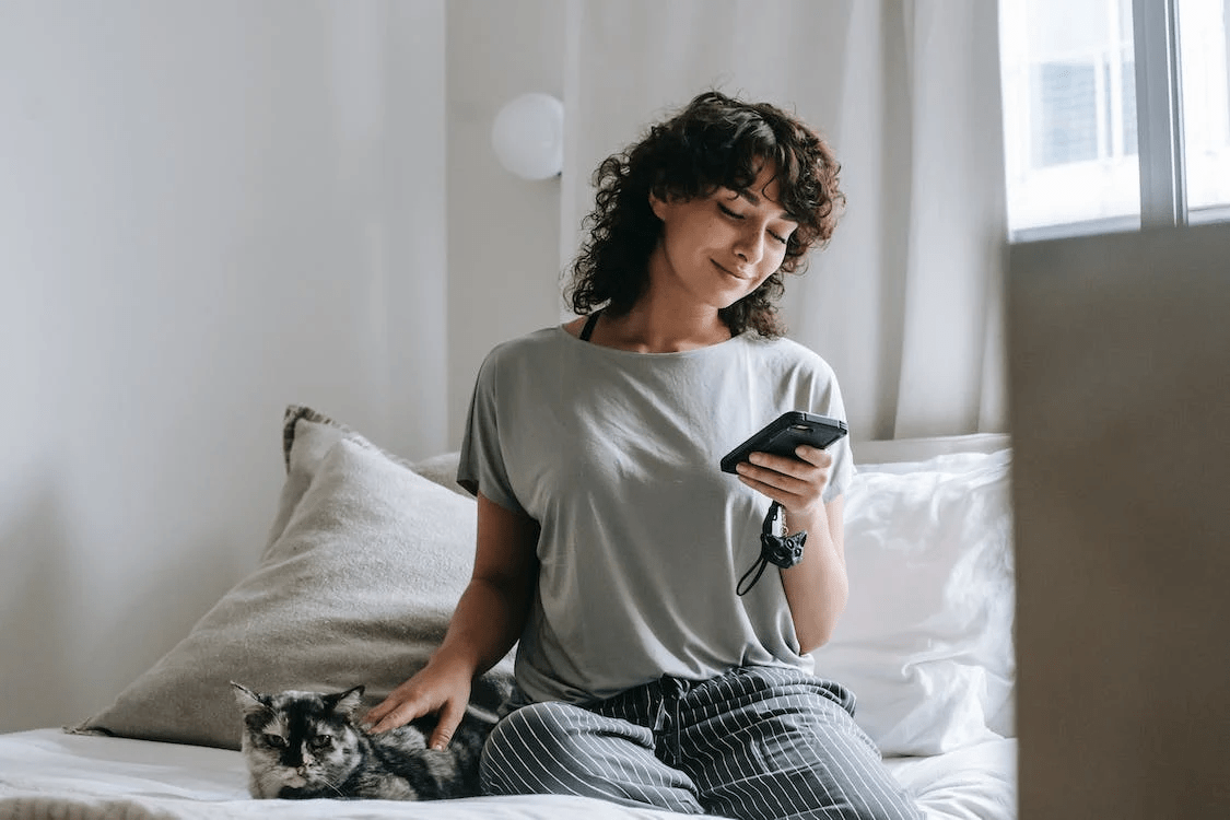 smiling young female messaging on smartphone and petting cat on bed
