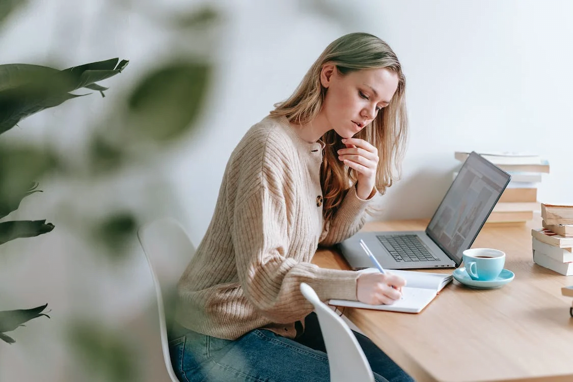 a woman taking notes at table with laptop and cup of coffee
