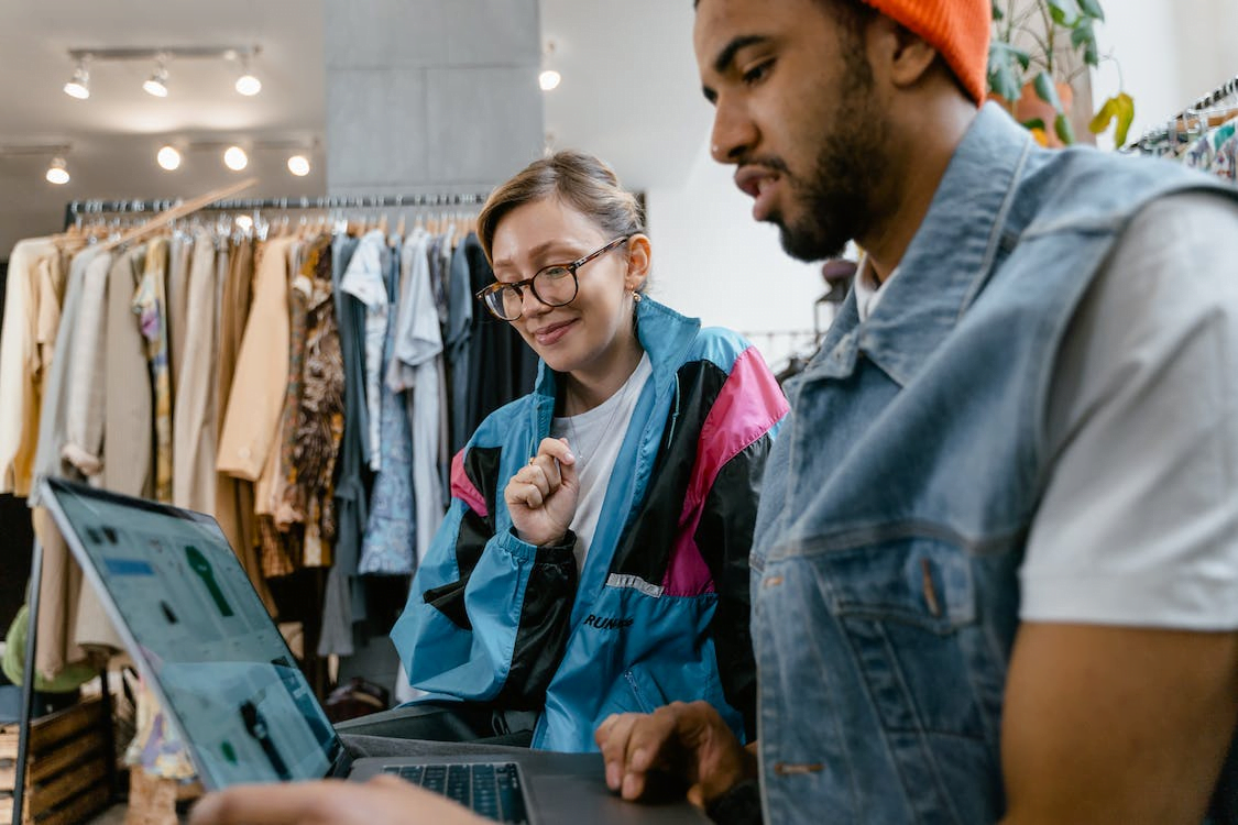 a man and a woman looking at a laptop screen with clothes on the background